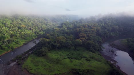 Misty-clouds-shroud-mountain-ridge-and-bend-in-fast-flowing-tropical-river,-aerial-reverse-dolly
