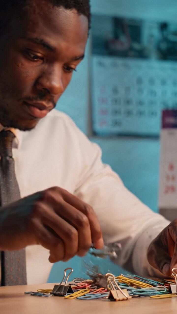 Concentrated Young Man Organizing Colorful Paper Clips on a Wooden Desk Surrounded by Office Supplies, Highlighting Focus and Attention to Detail in a Workspace
