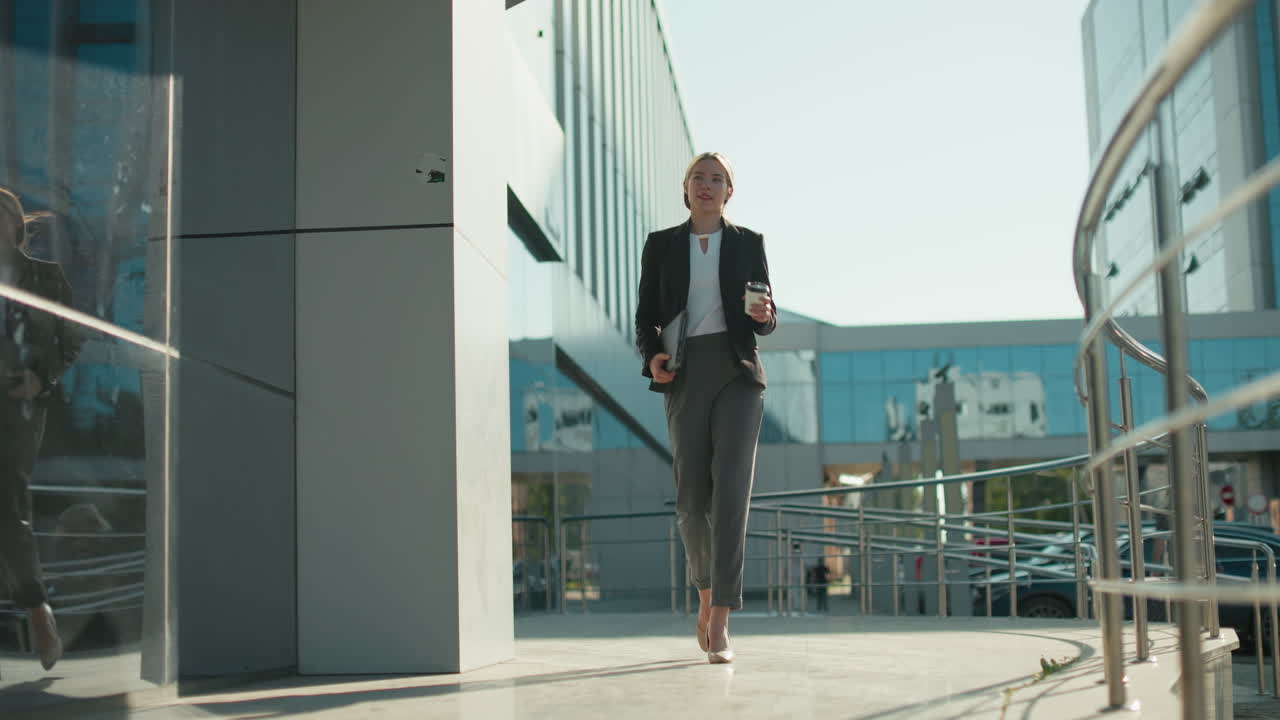 Confident woman in heels holding ipad and coffee walks outside modern building with mirrored windows reflecting her motion and parked cars, as another person walks in background