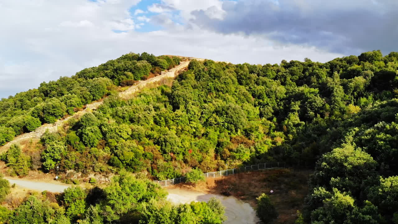 GREECE, ASPROVALTA - SEPTEMBER 26, 2020: Unpaved road with parked car, hill covered with lush greenery. Remains of ancient structures on the top on the hill