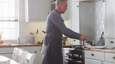 happy caucasian man wearing apron, standing in kitchen, cooking dinner