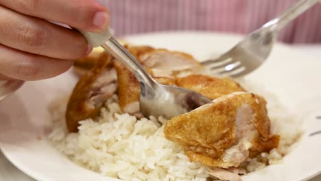 a hand uses a fork and spoon to lift a piece of roasted hainanese chicken from a plate of steamed rice under soft indoor lighting, captured in a medium close-up