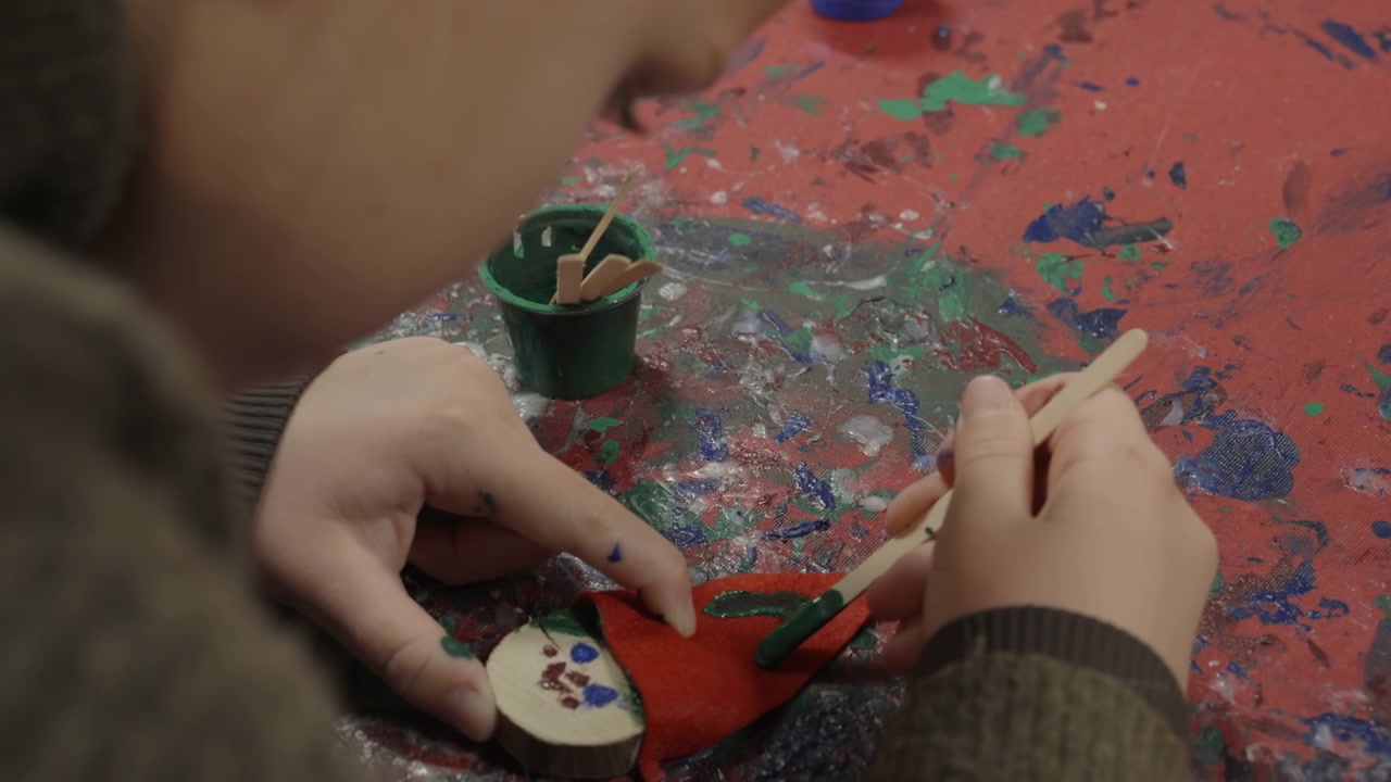 Primo piano delle mani di un bambino che dipingono motivi verdi su una piccola bambola fatta a mano di feltro e legno su un tavolo disordinato coperto di schizzi di vernice, mentre crea un lavoretto
