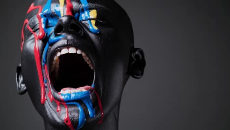 dark haired model wearing black makeup, multicolored paint dripping across face, screaming with wide open mouth against shadowy background, embodying dramatic artistic expression