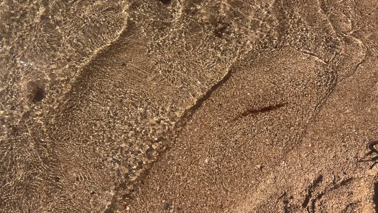 Close-up of transparent water gently washing over fine sandy beach, creating delicate ripples in Crete, Greece