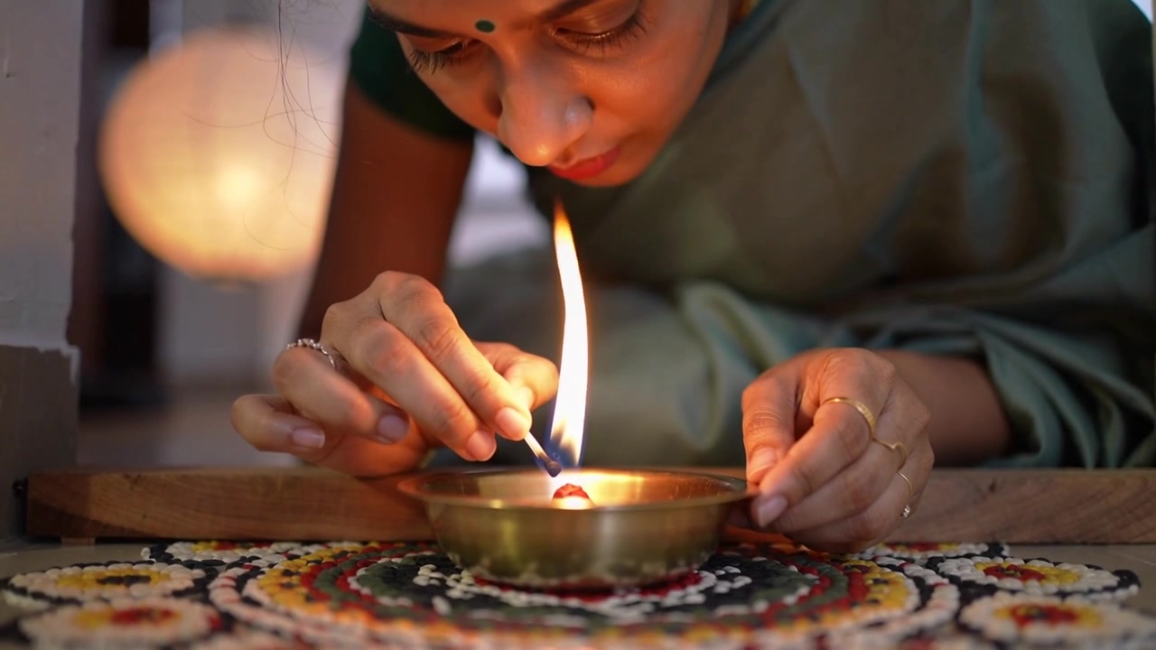 Lighting woman in green sari for prayer, leaning through doorway, steadying brass diya on rangoli