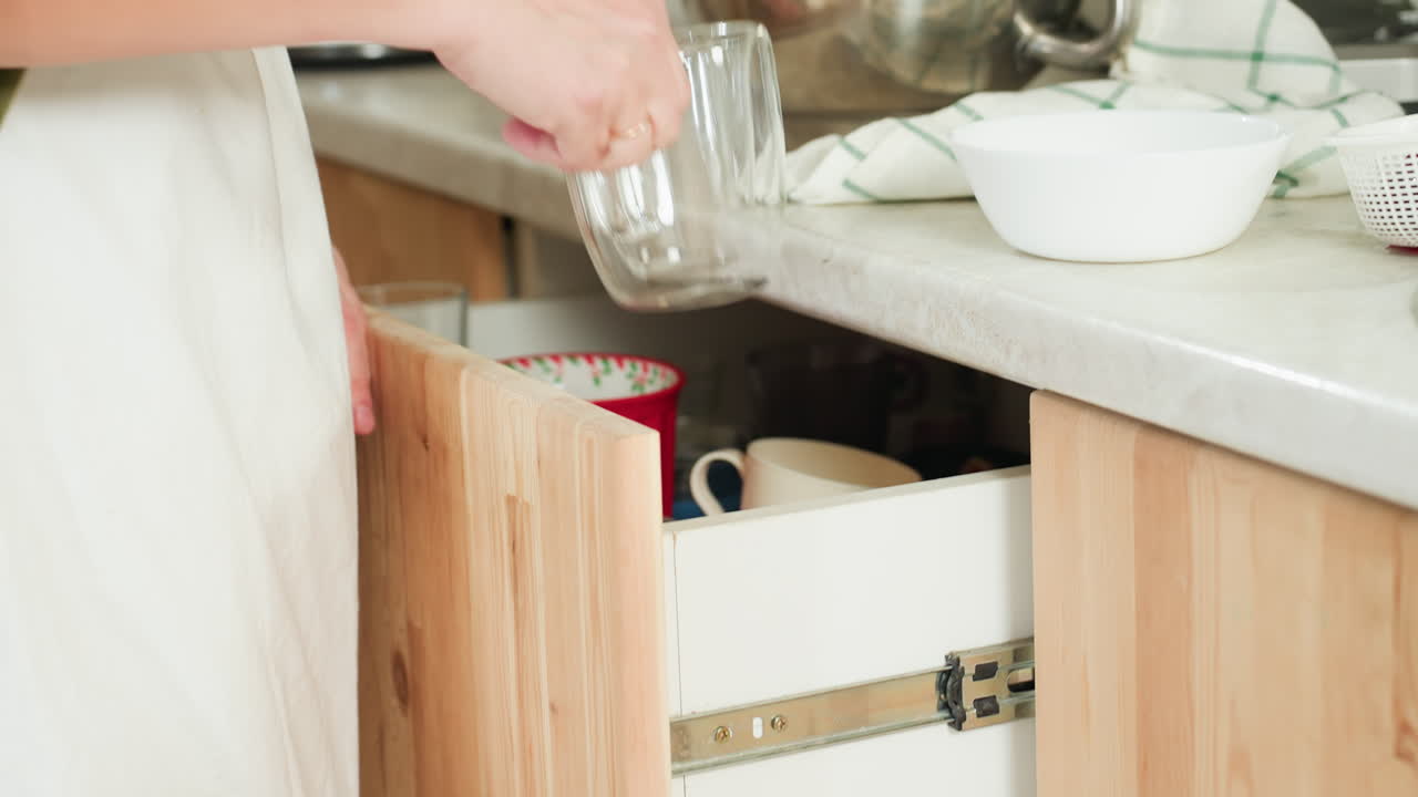 Close up of woman in kitchen retrieving glass cup from wooden drawer, placing it on countertop beside clean dishes and tea towel, then gently closing drawer
