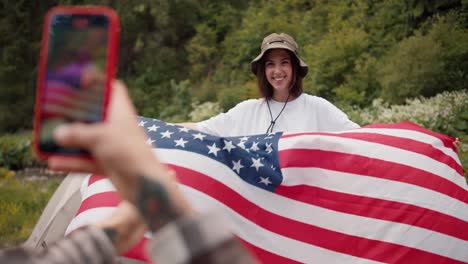 Shooting-close-up-a-girl-takes-a-picture-of-her-girlfriend,-a-brunette-girl-with-an-American-flag-in-her-hands,-which-is-fluttering-in-the-wind.-Then-she-shows-a-photo-that-she-took-against-the-backdrop-of-a-green-forest