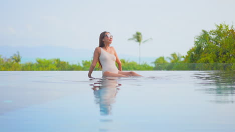 Slim-beauitufl-woman-sitting-in-the-shallow-swimming-pool-water,-sunbathing-during-hot-sunny-day-with-blurred-tropical-scenery-in-the-background