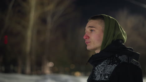 side view of young man wearing green hoodie and patterned winter jacket walking in cold night air, visible cold breath escaping from mouth, with soft bokeh lights and bare winter trees in background