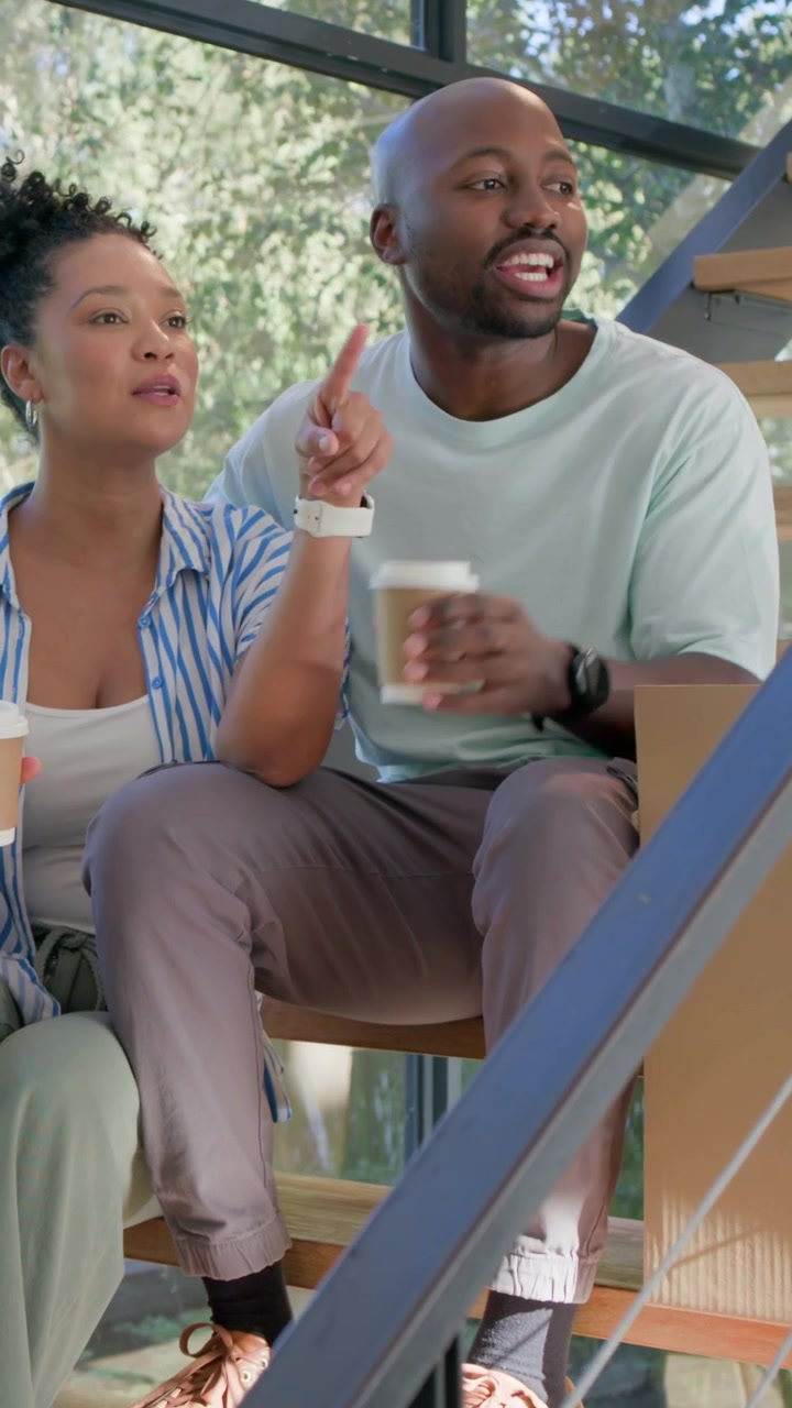 Vertical video: Sitting pair sipping coffee pointing at windows placing cup on box at home mid-move