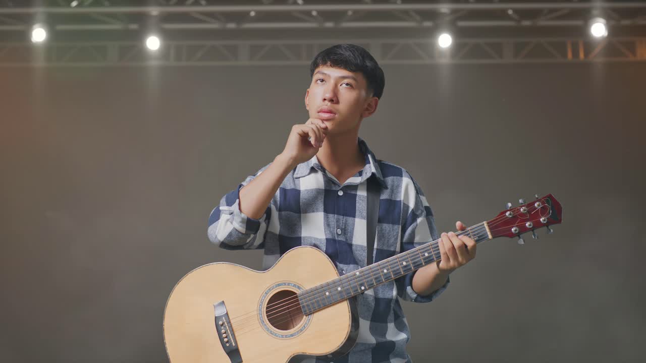 기타를 들고 있는 아시아인 십대가 무대에서 무언가를 생각하고 둘러보고 있다.