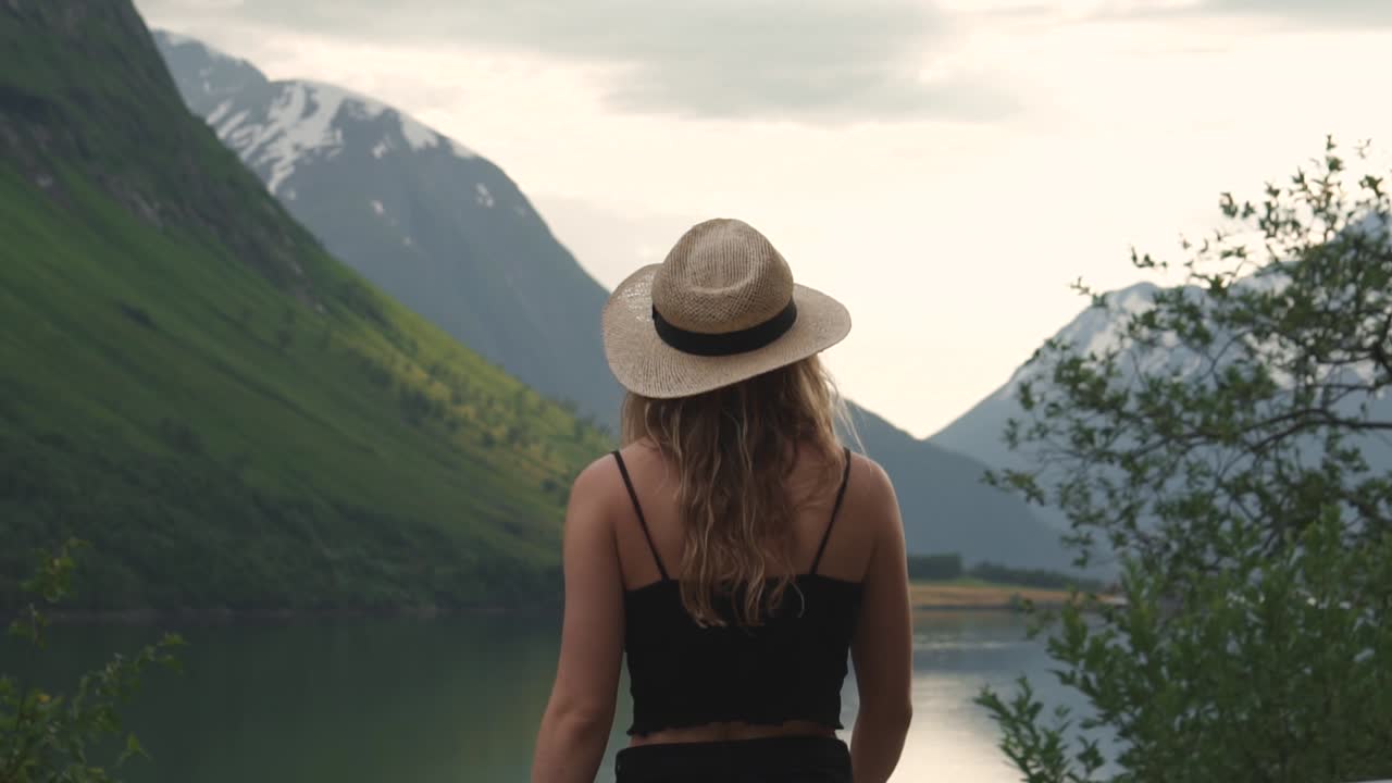 Slow motion shot of a girl wearing a hat, walking towards a lake, taking in the relaxing environment, surrounded by hills and nature.