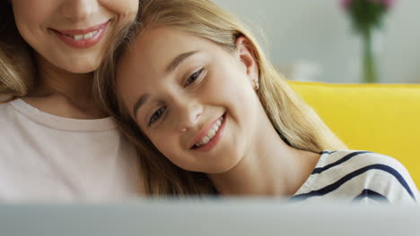 close up of the cute teenage girl leaning on her mother's shoulder while watching something on laptop in living room