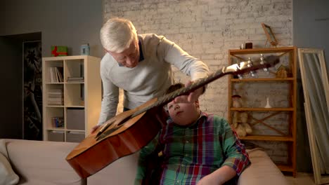 grandpa gives his grandson a guitar, a gift, a surprise. an elderly man gives a guitar to a young fat child. home comfort, family idyll, cosiness concept, difference of generations, close up 60 fps