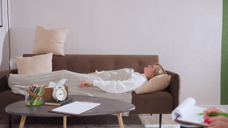 visitor lies straight on sofa and wraps blanket around herself in cozy therapy space next to table with clock pencils notebook creating warm healing atmosphere for intimate counseling session