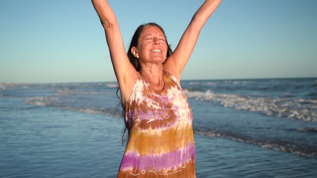 Woman In Her 50s Raising Arms With Eyes Closed. Breathing Exercise At The Beach On A Sunny Morning. medium shot
