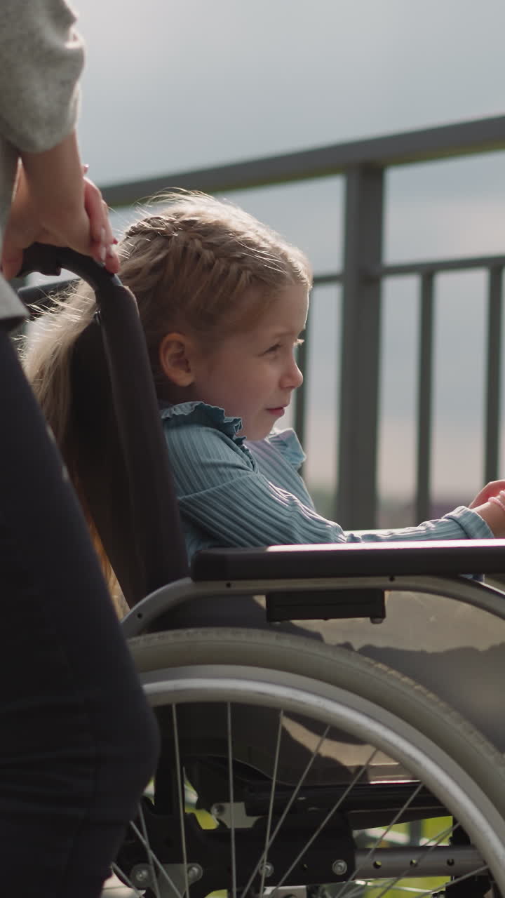 una niña pequeña se sienta en una silla de ruedas señalando con el dedo a una torre alta. madre e hija con lesión en la médula espinal disfrutan de la vista de la ciudad desde la plataforma de observación