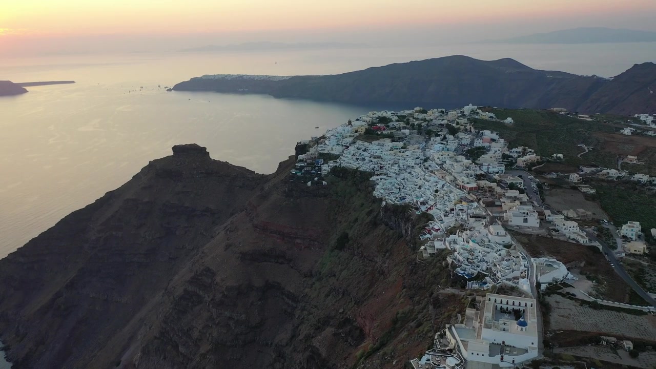 Hisnande vidvinkel drönarbild av Fira, Santorini, i skymningen. Visar traditionella vita byar byggda längs massiva vulkaniska klippor med utsikt över lyxyachter ankrade i det lugna Medelhavet.