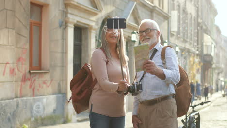 Senior-Couple-Of-Tourists-Standing-At-The-Sunny-Street-Of-The-Nice-Town-And-Taking-Selfie-Photos-With-Smartphone-Camera-On-The-Selfie-Stick