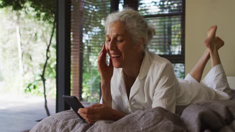 Ältere-Frau-Mit-Gemischter-Abstammung-Liegt-Mit-Smartphone-Im-Bett-Und-Lächelt