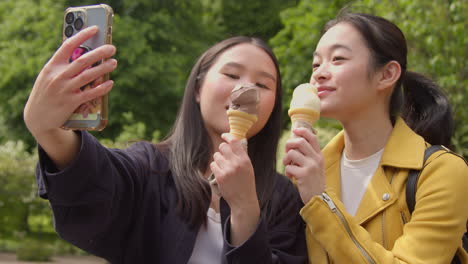 Two-Smiling-Young-Female-Friends-Meeting-And-Posing-For-Selfie-Eating-Ice-Cream-Outdoors-In-Park-Together