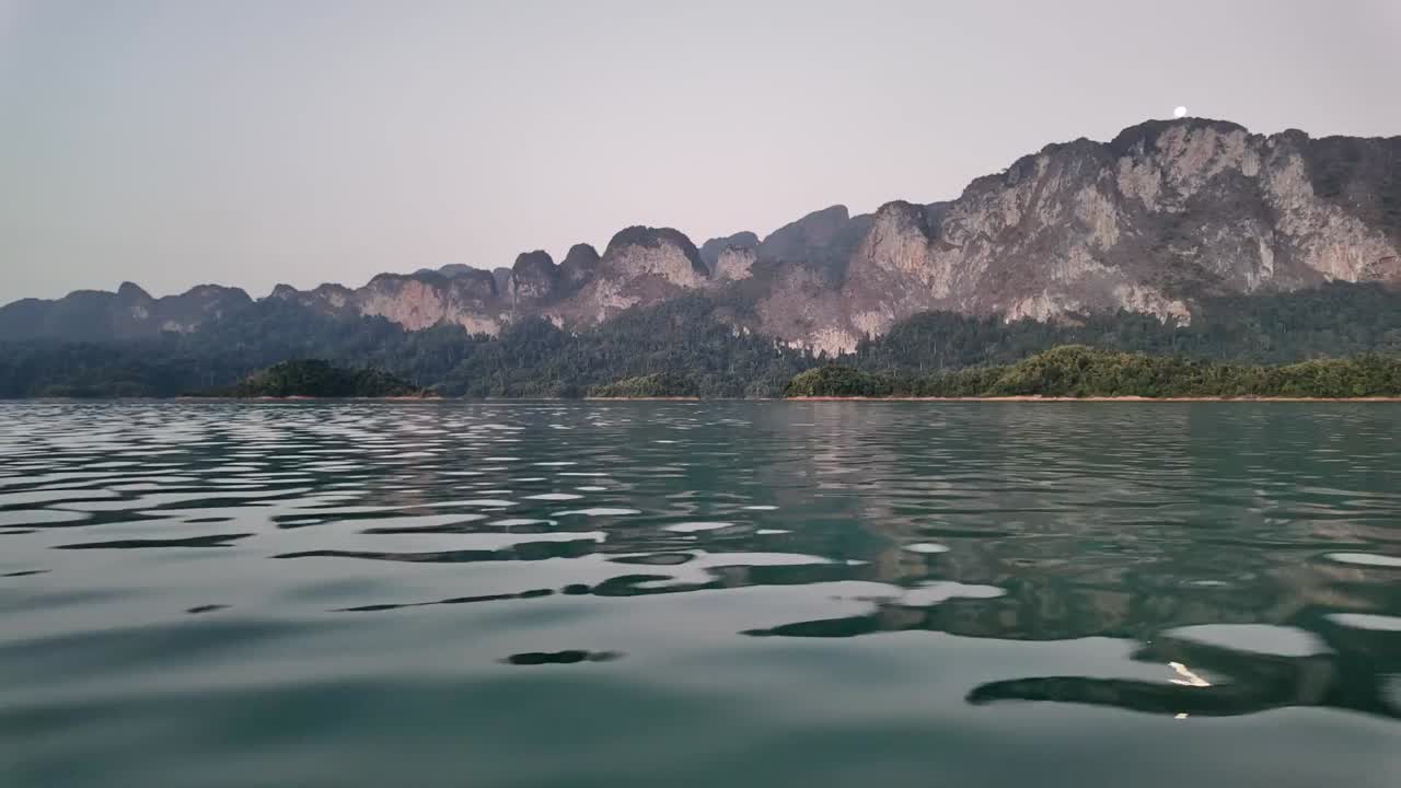 панорамный клип, показывающий традиционную деревянную лодку, движущуюся по мелководью, окруженную красивым горным и лесным ландшафтом