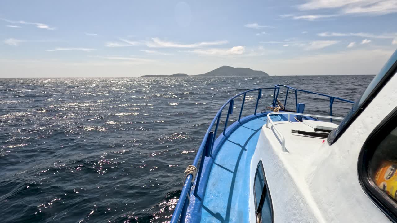 Blue boat moves toward distant island on sparkling sea, daylight, steady camera, clear sky