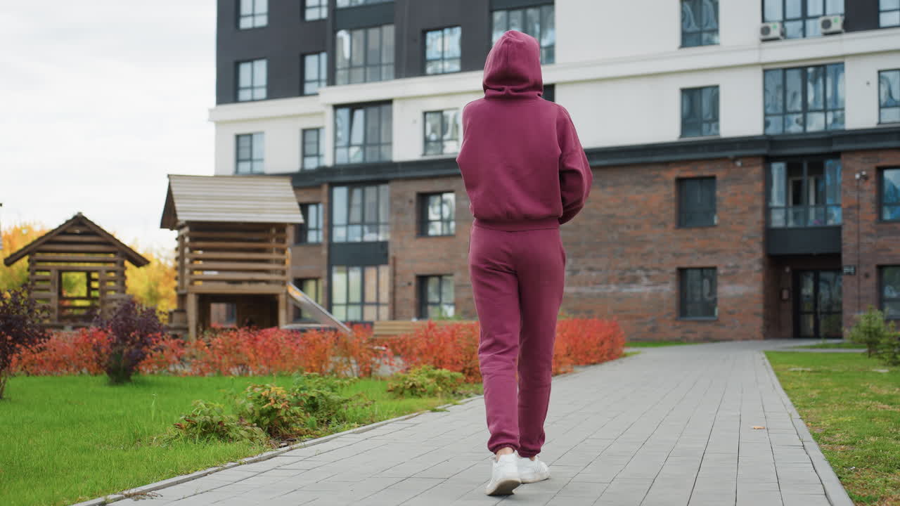 Back view of resilient woman in maroon hoodie walking toward modern urban building past decorative wooden birdhouses beside paved path lined with spring shrubbery and fresh green grass
