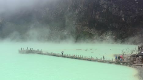 aerial drone of a green toxic sulfur lake inside a volcano called kawah putih with fumes and gasses rising while tourists walk a long bridge in bandung indonesia