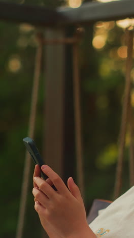 young girl types messages holding smartphone on terrace. toddler relaxes in cozy armchair surrounded by woodland area. calm child rests alone at sunset