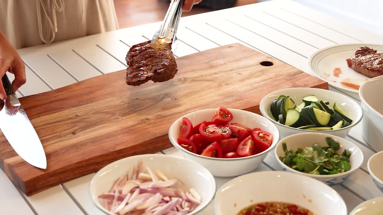 A person slices a steak on a wooden board surrounded by fresh vegetables in a bright kitchen