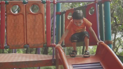 A-young-boy,-wearing-a-Santa-face-mask,-joyfully-slides-down-a-colorful-roller-slide-on-a-tropical-beach-playground