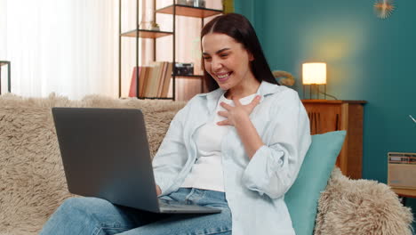 young woman on sofa watching funny video on laptop screen laughing joyfully at amusing content