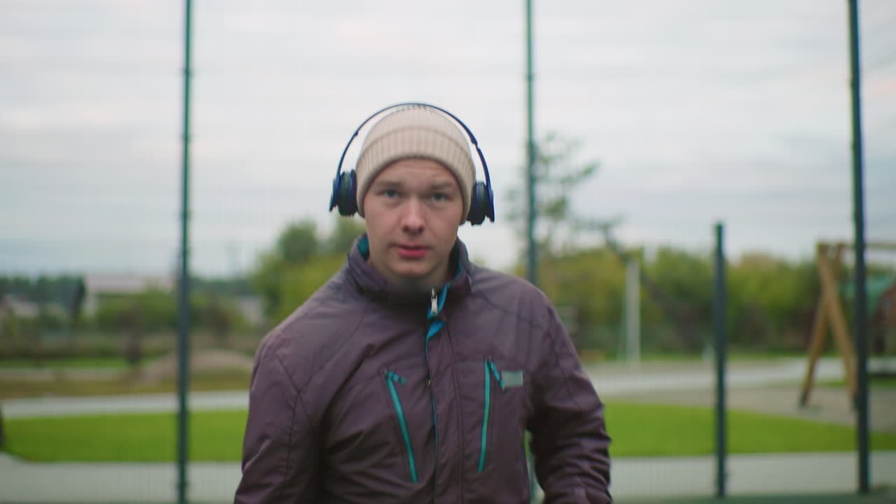 Mann utendørs med hodetelefoner som snakker direkte til kameraet på inngjerdet parkbane, iført lue og jakke, rolig overskyet stemning, avslappet veiledningstone, subtil pust synlig, hender som gestikulerer, livsstil