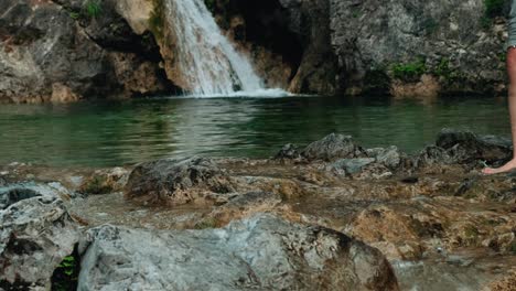 Hiker-walks-across-a-rocky-stream-barefoot-with-beautiful-waterfall-on-the-background-in-slow-motion