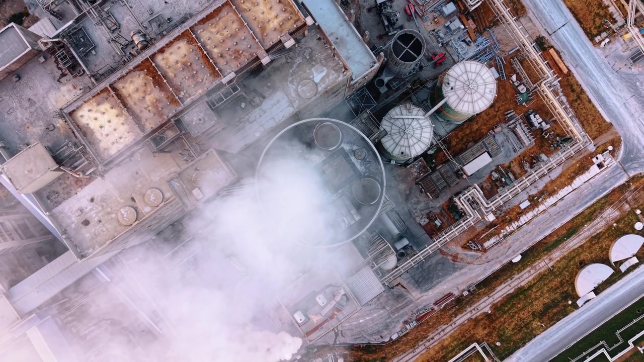 Top view of industrial tower releasing dense steam. Vertical drone perspective shows a circular stack, tanks and pipe grids beneath drifting white vapor.