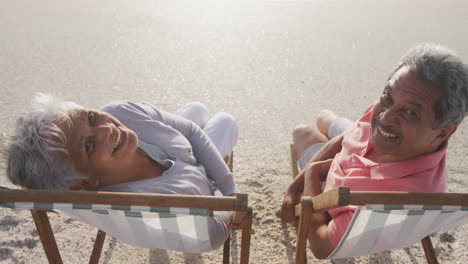 feliz pareja hispana senior relajándose en tumbonas en la playa al atardecer y mirando a la cámara