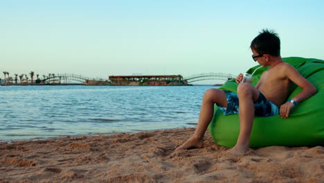 young man drinking juice at coastline. teenager sitting in chair bag at beach.