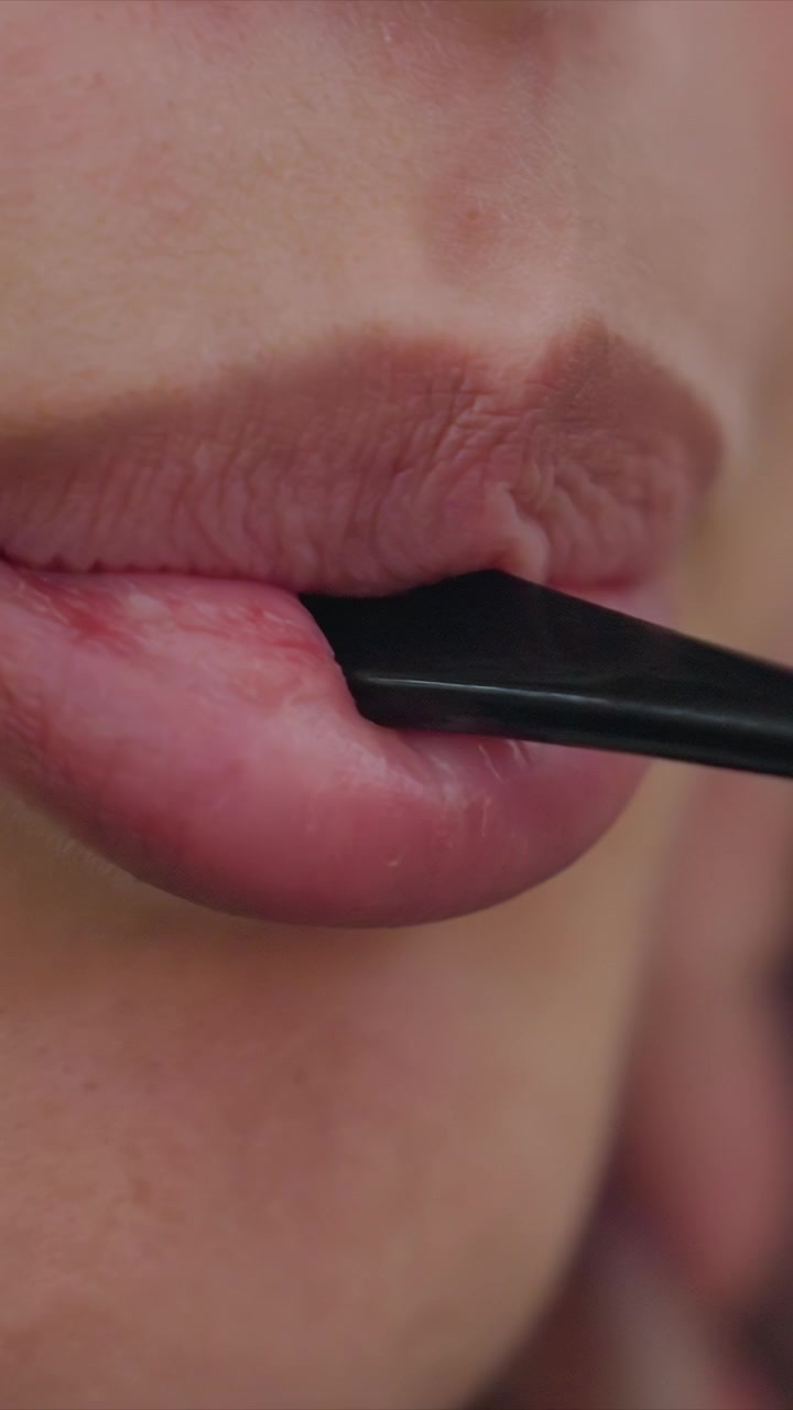 Close up of lips applying lipstick with black applicator. Applicator moves with slight motion for precision. Setting appears minimal with emphasis on detailed application