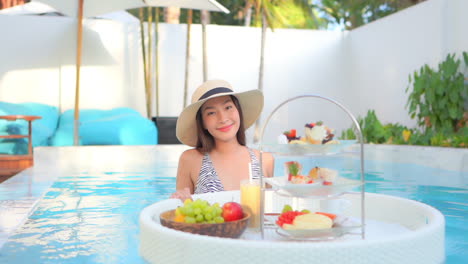 a pretty young woman in a swimming pool pushes a floating banquet of fruit and desserts toward the camera