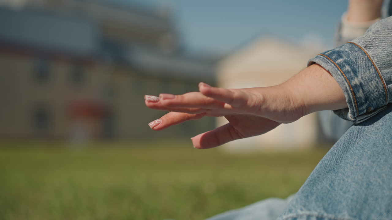 Hand streicht über Gras in einem Vorstadtpark, Person in Jeansjacke, Finger streichen über Grashalme unter warmem Nachmittagslicht, verschwommene Häuser im Hintergrund, langsame, filmische Bewegung, Schriftsteller pausiert.