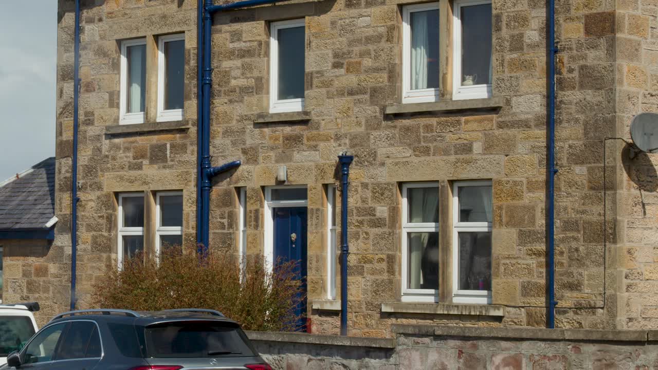 Camera pans upward on stone house exterior, parked cars, overcast sky, Cromarty, Scotland