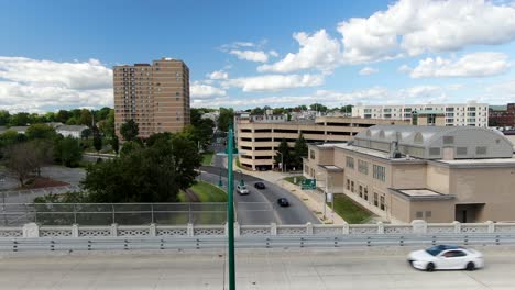 Cars-pass-by-on-overpass-bridge,-traffic-on-beautiful-summer-day,-rising-aerial-reveals-American-USA-city,-decorative-fancy-green-light-lamp-fixture,-establishing-drone-shot