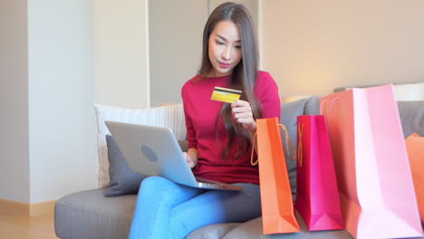 una chica asiática sentada en el sofá rodeada de bolsas de compras escribe la información de la tarjeta de crédito en su cuaderno en casa