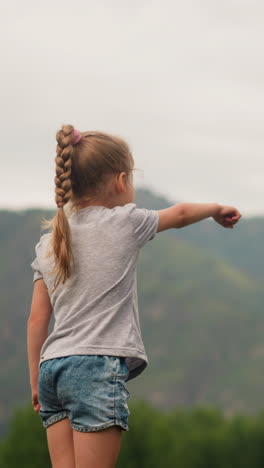 cute playful little child girl with braided hair points to large distant mountains at highland resort on summer day backside view slow motion