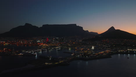 Increíble-Toma-Aérea-De-La-Ciudad-Al-Atardecer.-Marina-Y-Calles-Y-Edificios-Iluminados-En-El-Barrio-Costero.-Silueta-De-La-Cresta-De-La-Montaña-En-El-Fondo.-Ciudad-Del-Cabo,-Sudáfrica