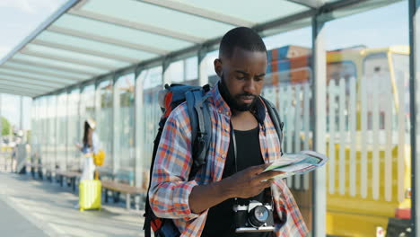 joven viajero afroamericano con mochila sosteniendo un mapa de la ciudad mientras camina en la estación de autobuses