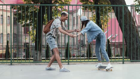 full length view of a teenage girl riding a skateboard while her african american boyfriend helping her maintain balance in the street