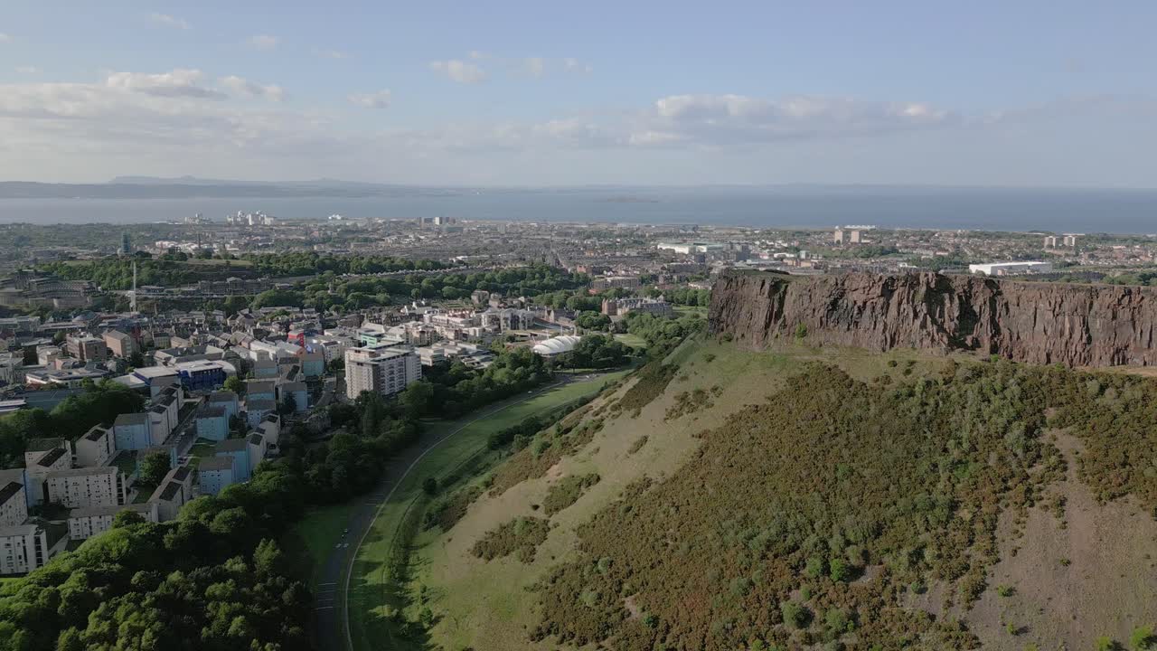 Aerial push in of Salisbury Crags with overlooking urban Edinburgh city centre in background, Edinburgh, Scotland, UK